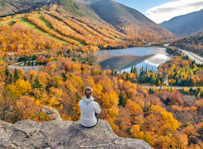 Beautiful fall photo of young woman sitting on rock bluff overlooking small valley lake.